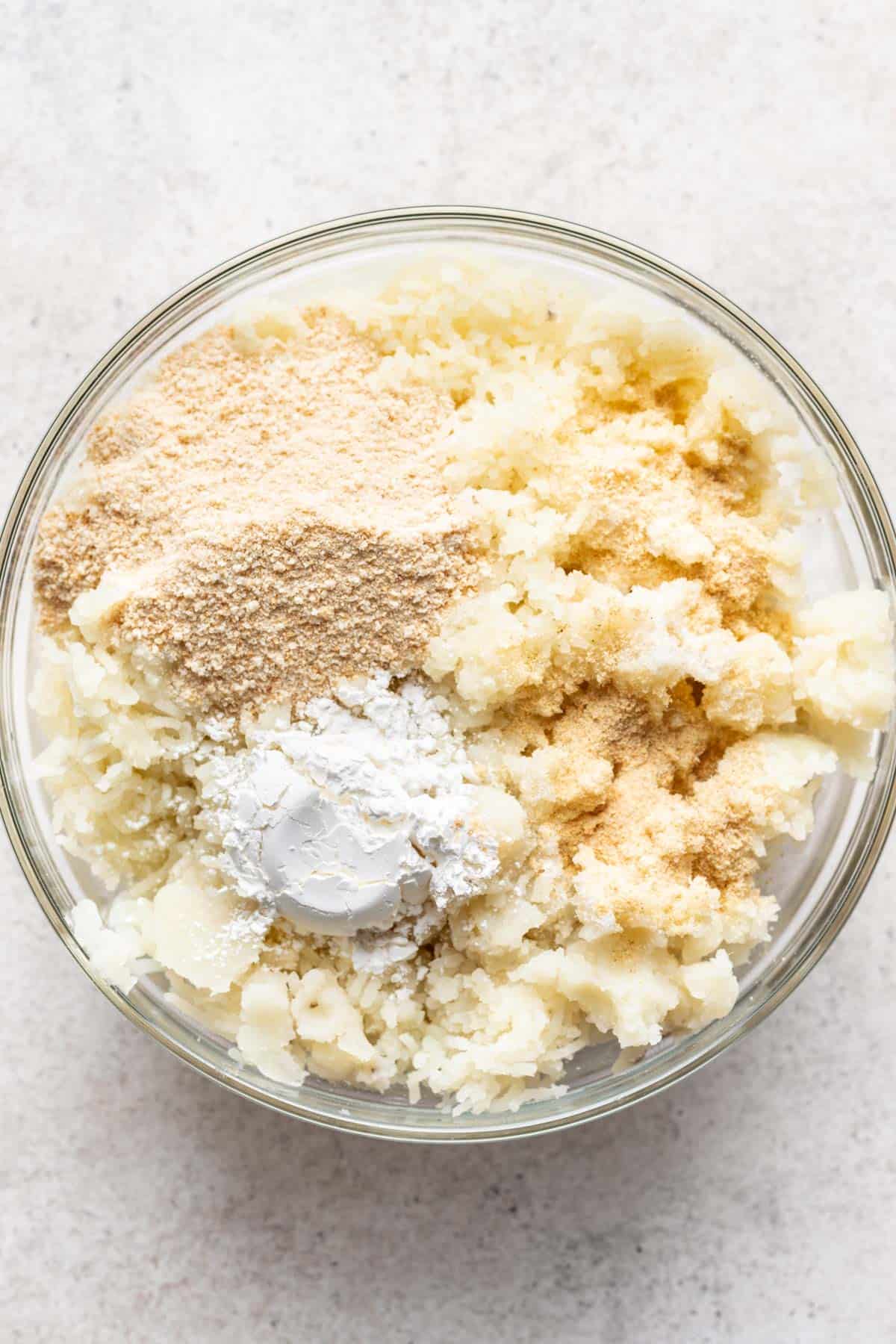 Breadcrumbs, cornstarch, and seasonings added to mashed potatoes in a glass bowl.
