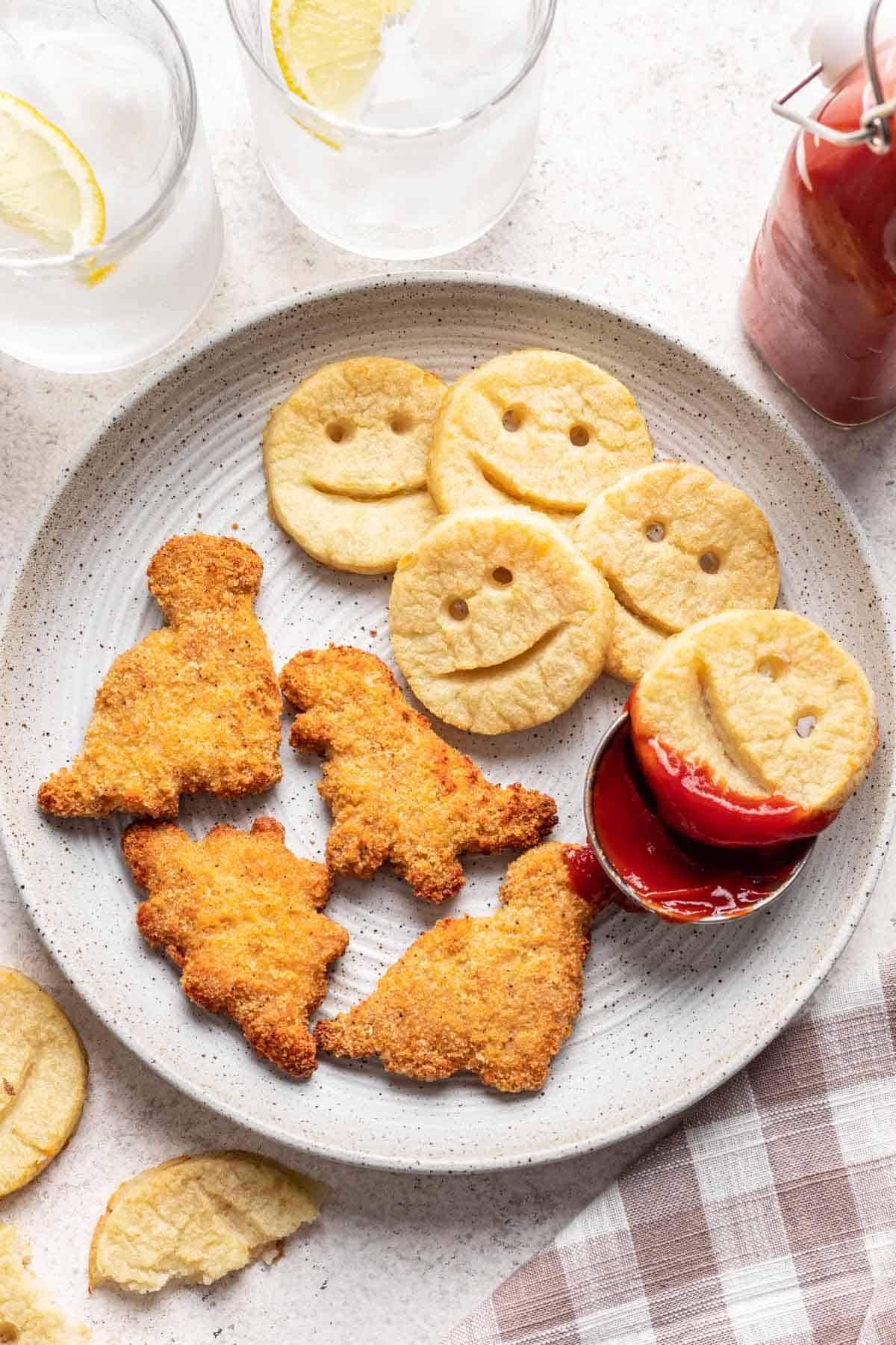 Smiley potatoes on a plate with homemade dinosaur chicken nuggets and a side of ketchup.