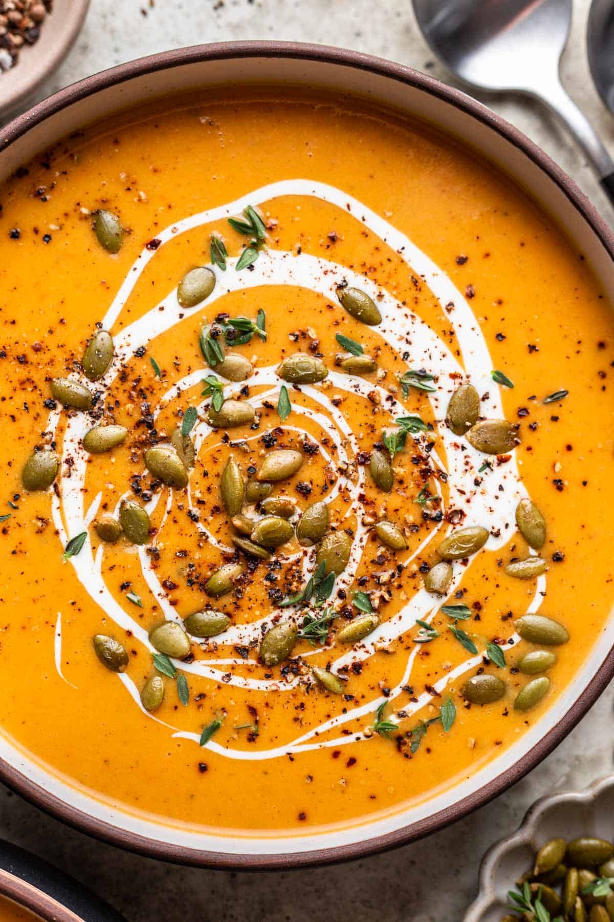 Butternut squash and carrot soup in a bowl with fresh thyme, pumpkin seeds, cream, and cracked pepper on top.