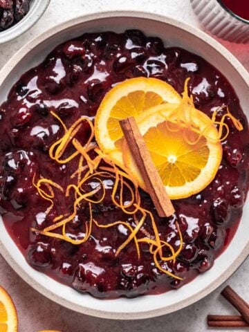 Cranberry sauce with dried cranberries in a bowl, topped with sliced oranges and a cinnamon stick.