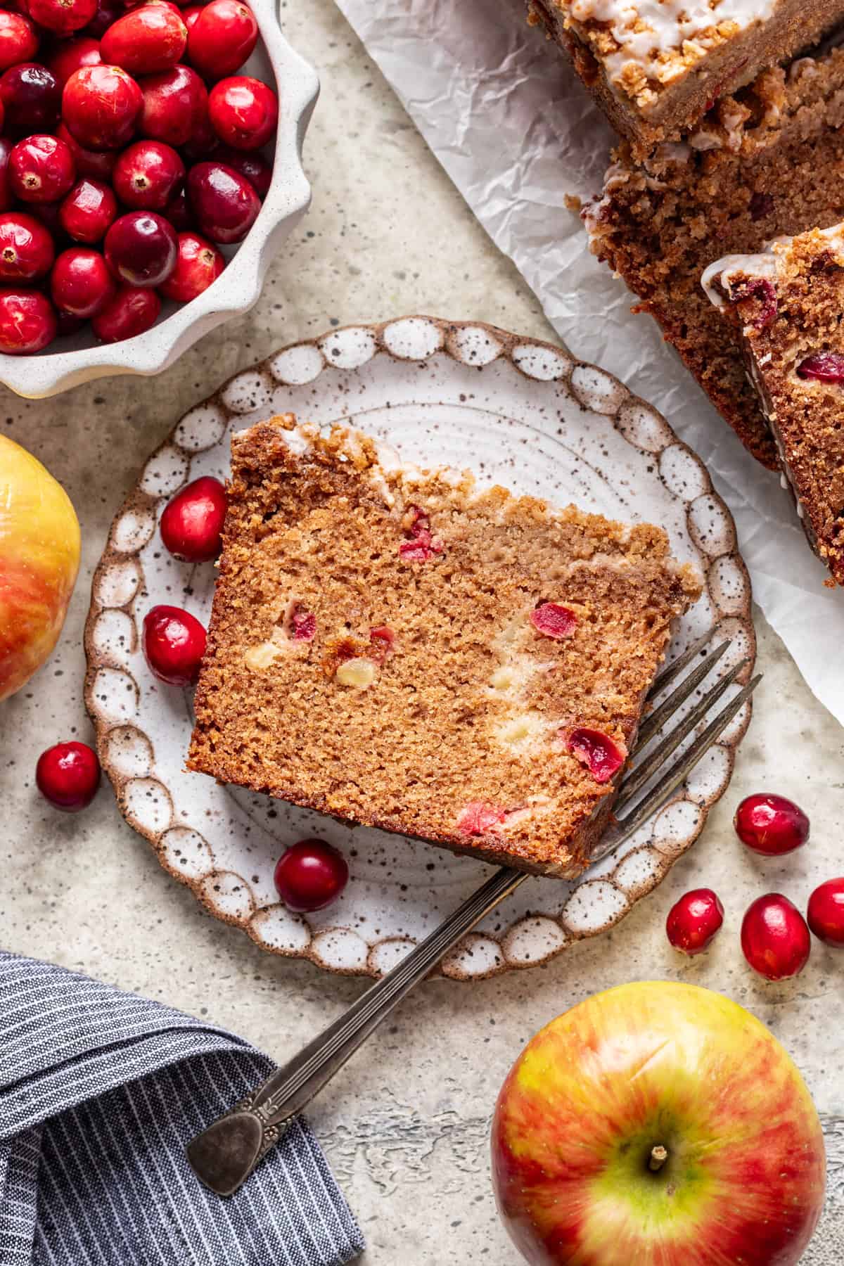 A slice of cranberry apple bread on a plate with fresh cranberries and apples around it.