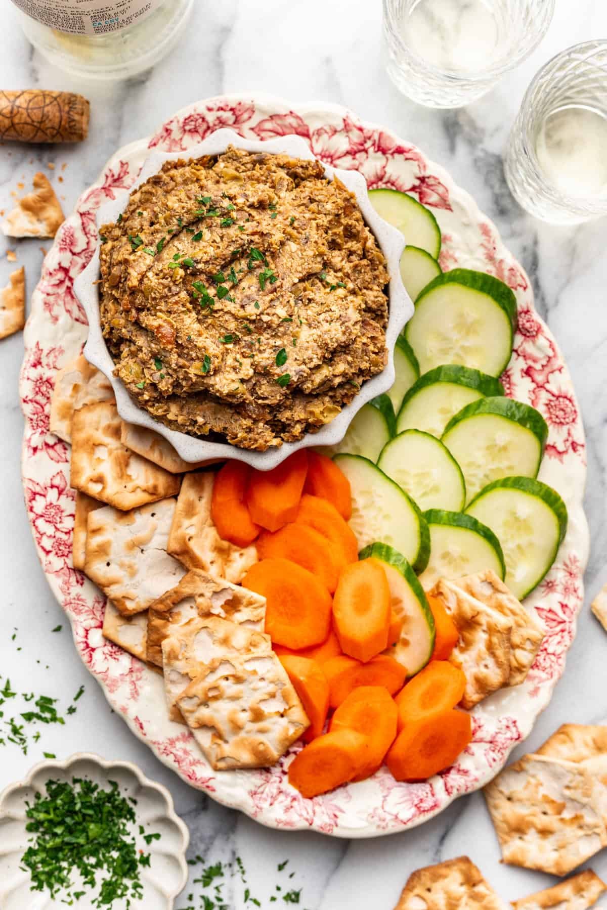 Vegetarian chopped liver in a serving bowl with sliced cucumbers, carrots, and crackers on the side.